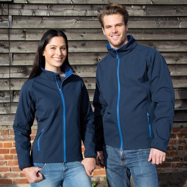 Klassisk herresoftshell sæt To personer står ved siden af hinanden iført blå jakker og jeans, smilende, foran en trævæg på en solrig dag.