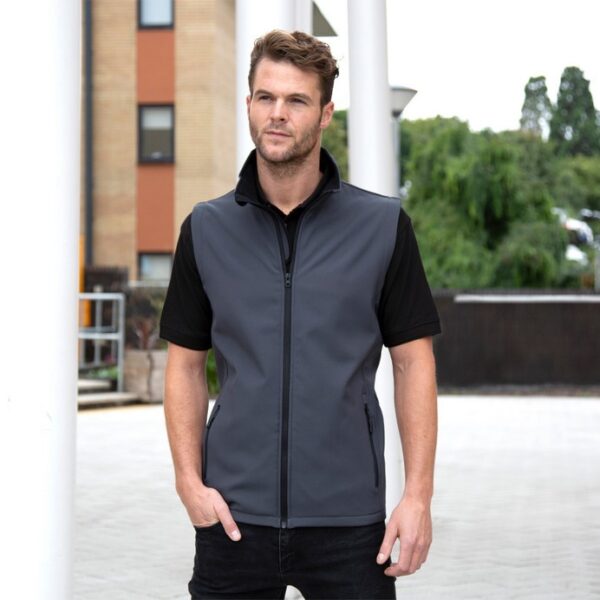 Klassisk herresoftshell vest En mand iført en sort t-shirt og grå vest står afslappet med hænderne i lommerne foran en bygning med grønne buske i baggrunden.