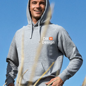 Mand iført grå hættetrøje står smilende. Tekst på trøjen: "Dit Design." Blå himmel og tørre kornstilke i baggrunden.