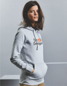Kvinde iført en grå hættetrøje med teksten "D+ Design" står afslappet med hænderne i lommerne i en neutral, lys grå omgivelse.