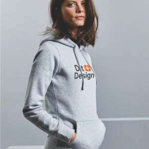 Kvinde iført en grå hættetrøje med teksten "D+ Design" står afslappet med hænderne i lommerne i en neutral, lys grå omgivelse.