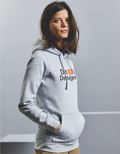 Kvinde iført en grå hættetrøje med teksten "D+ Design" står afslappet med hænderne i lommerne i en neutral, lys grå omgivelse.