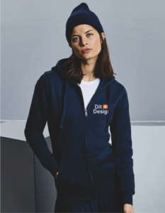 Kvinde står iført mørkeblå hættetrøje med teksten "Dit Design" og en rød-hvid firkant, i minimalistisk indendørs miljø med lys baggrund. Hun bærer også en hue og T-shirt.