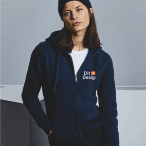 Kvinde står iført mørkeblå hættetrøje med teksten "Dit Design" og en rød-hvid firkant, i minimalistisk indendørs miljø med lys baggrund. Hun bærer også en hue og T-shirt.
