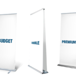 Tre reklamebannere står ved siden af hinanden med tekst på hvert: "BUDGET" på det venstre banner, "DOUBLE" på det midterste, og "PREMIUM" på det højre. Baggrund er grå med skygger.