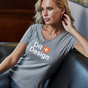 Kvinde iført en grå T-shirt sidder, læner sig mod en sofa. T-shirten har teksten "Dit Design" med et orange plustegn. Omgivelserne er et elegant indendørs rum.