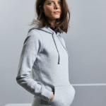 En person med langt, mørkt hår står i profil og har hænderne i lommerne på en grå hættetrøje. Baggrunden er minimalistisk og neutralt lys grå.