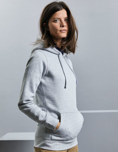 En person med langt, mørkt hår står i profil og har hænderne i lommerne på en grå hættetrøje. Baggrunden er minimalistisk og neutralt lys grå.
