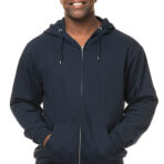 En mand iført en mørkeblå hættetrøje med hænderne i lommerne smiler foran en hvid baggrund. Han har lyseblå jeans på.