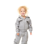 Et lille barn iført en grå træningsdragt står smilende og barfodet på en hvid baggrund, med krøllet hår og armene afslappet ved siderne.