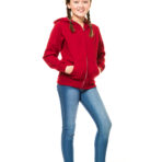 En pige iført en rød hættetrøje og blå jeans står med hænderne i lommerne og smiler. Baggrunden er hvid og enkel uden andre elementer.