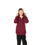 Et barn trækker op i hætten af sin bordeauxrøde hættetrøje og smiler. Barnet har sorte bukser og sorte sneakers på og står mod en hvid baggrund.