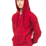 En ung person iført en rød hættetrøje står afslappet med hænderne i lommerne. Personen har lyst hår og kigger fremad. Baggrunden er hvid og neutral.