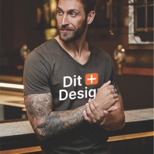 Mand bærer en mørk T-shirt med teksten "Dit Design" trykt på forsiden. Han står ved en bardisk i en stilfuld indretning, med tatoverede arme synlige.
