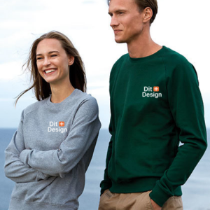 To personer står smilende, iført sweatshirts, på en udendørs, blæsende kyst. Ord på tøj: "Dit Design". Den ene trøje er grå, den anden er grøn.