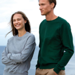 To personer iført sweatshirts står sammen, en kvinde i grå og en mand i grøn. De smiler ved kysten med havet i baggrunden.