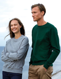 To personer iført sweatshirts står sammen, en kvinde i grå og en mand i grøn. De smiler ved kysten med havet i baggrunden.
