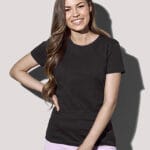 En kvinde iført en sort t-shirt og lyserøde shorts smiler, står med hånden på hoften og den anden foran sig, mod en ensfarvet grå baggrund.