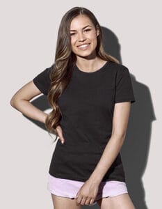 En kvinde iført en sort t-shirt og lyserøde shorts smiler, står med hånden på hoften og den anden foran sig, mod en ensfarvet grå baggrund.