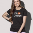 Kvinde står smilende iført sort t-shirt med teksten "Dit Design" i hvidt og orange. Hun har langt, mørkt hår og står mod en lys baggrund.