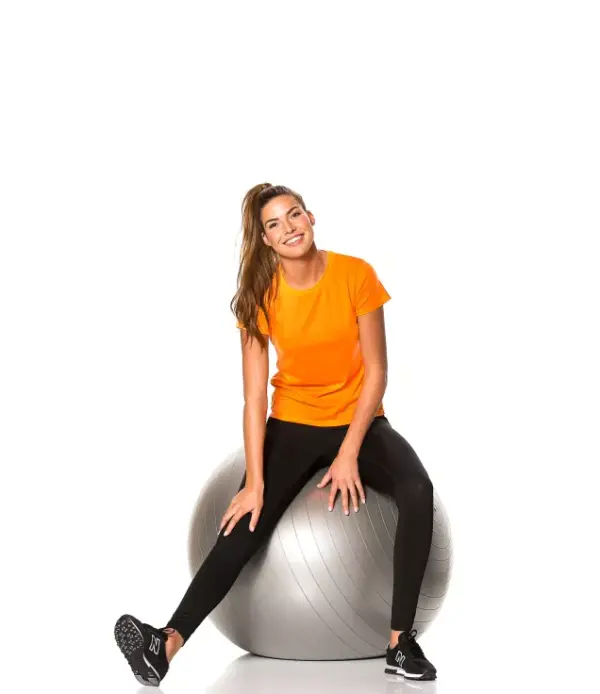 000250_Lady_Go_Sport_01.jpg Person sidder på en træningsbold iført en orange t-shirt og sorte bukser. Personen ser fremad i et hvidt, neutralt rum.
