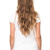 En person står med ryggen til, iført en hvid t-shirt og sorte bukser, med langt, bølget hår i en hvid baggrund.
