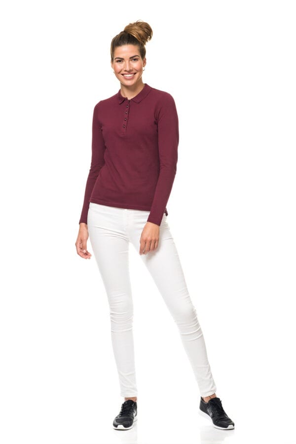 000810_Lady_Stretch_Polo_LS_01_1 En kvinde iført en bordeaux langærmet trøje og hvide bukser, står og smiler foran en hvid baggrund. Hun har sorte sneakers på og hendes hår er sat op i en knold.