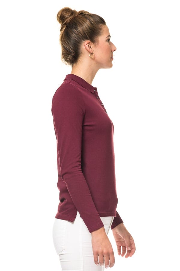 000810_Lady_Stretch_Polo_LS_02c_1 En kvinde står i profil, klædt i en bordeaux langærmet trøje og hvide bukser. Hendes hår er bundet op i en knold, og baggrunden er hvid.
