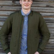 En mand smiler og poserer med hænderne på hofterne iført en olivengrøn jakke og en blå skjorte foran en væg af vejrbidt træ.