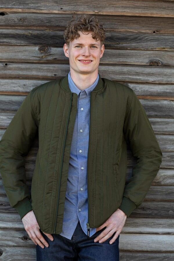 0886_570_image_1 En mand smiler og poserer med hænderne på hofterne iført en olivengrøn jakke og en blå skjorte foran en væg af vejrbidt træ.