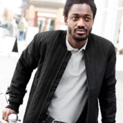 Manden står med hænderne på et cykelstyr i en bymiljø. Han har en sort jakke og grå polo på, bygninger kan ses i baggrunden.