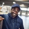Mand iført blå vest og kasket smiler, læner sig mod en papkasse i en oplyst industrihal.