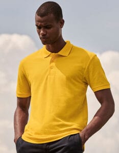 En mand iført en gul poloshirt står med hænderne i lommerne på et par blå bukser. Han ser nedad. Baggrunden består af skyer og en lys himmel.