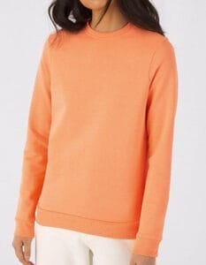 En person iført en lys orange sweatshirt står oprejst i front af et lyst, ensfarvet baggrund. Personen har hvide bukser på og har langt mørkt hår.