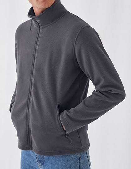 En person iført en mørkegrå hættetrøje står med hænderne i lommerne. Trøjen har lynlås foran og bæres i en neutral, minimalistisk stil på lys baggrund.