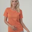 En kvinde iført en orange t-shirt og grå bukser står med den ene hånd på maven og ser direkte fremad. Baggrunden er ensfarvet og lys.