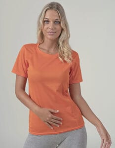 En kvinde iført en orange t-shirt og grå bukser står med den ene hånd på maven og ser direkte fremad. Baggrunden er ensfarvet og lys.