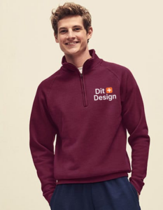 Ung mand iført vinrød trøje med lynlås og teksten "Dit Design" med et lille orange plus-tegn. Han smiler i et neutralt, lyst rum.