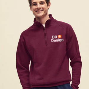 Ung mand iført vinrød trøje med lynlås og teksten "Dit Design" med et lille orange plus-tegn. Han smiler i et neutralt, lyst rum.