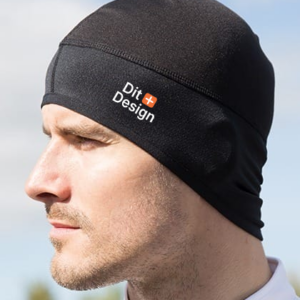 En person iført en sort hue, kigger til siden. Huen har teksten "Dit Design" trykt på siden. Baggrunden er sløret med blå himmel og grønne træer.