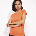 En kvinde iført en orange t-shirt står med krydsede arme i et studiemiljø med hvid baggrund.