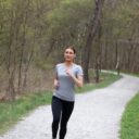 En kvinde løber på en grussti iført grå t-shirt og sorte leggings. Stien snor sig gennem en skov med træer og grøn vegetation omkring.