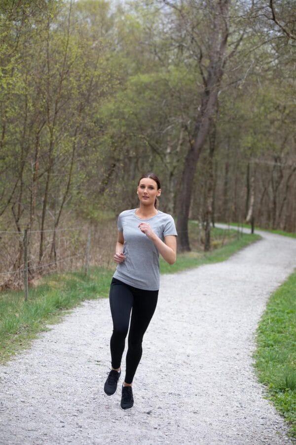 g11020_210_image_1 En kvinde løber på en grussti iført grå t-shirt og sorte leggings. Stien snor sig gennem en skov med træer og grøn vegetation omkring.