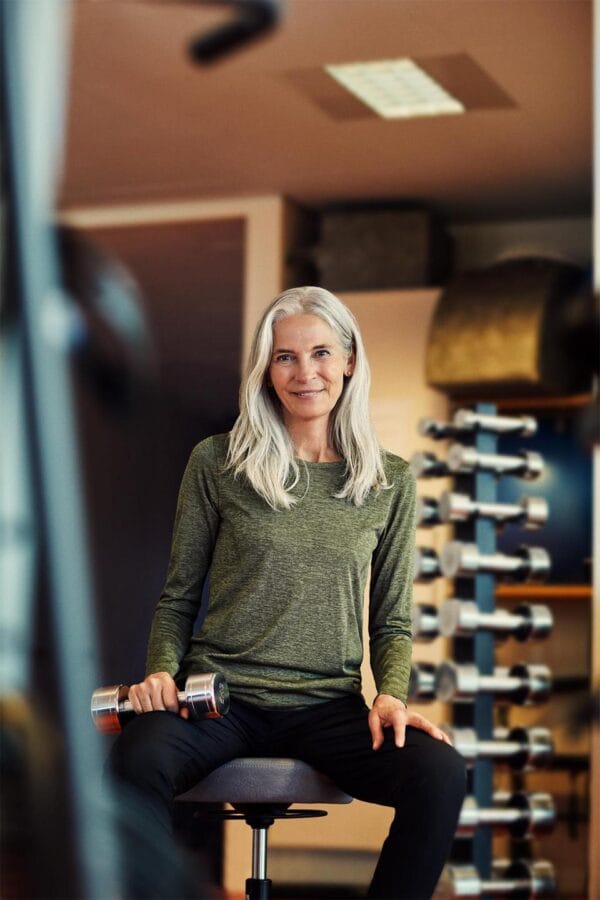 g11021_562_image_1 En kvinde med gråt hår sidder på en stol og holder en håndvægt i en træningssal omgivet af håndvægte og træningsredskaber.