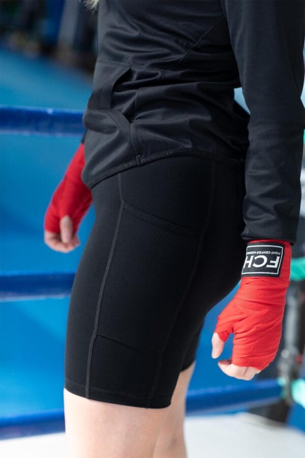 g11049_900_image_1 En person iført sorte træningstights og en sort trøje står i en boksering med røde håndbind. Der står "FCH" på håndbindet.