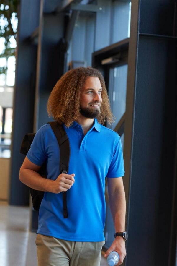 g21006_770_image_1 Manden med krøllet hår og blå skjorte bærer en rygsæk, mens han smilende går indendørs i et moderne bygning. Han holder en flaske i hånden.