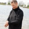 Mand i sort jakke står ved en sø og justerer sit ærme. I baggrunden er moderne hvide bygninger og grønne træer. Tekst på jakken: "Sport".