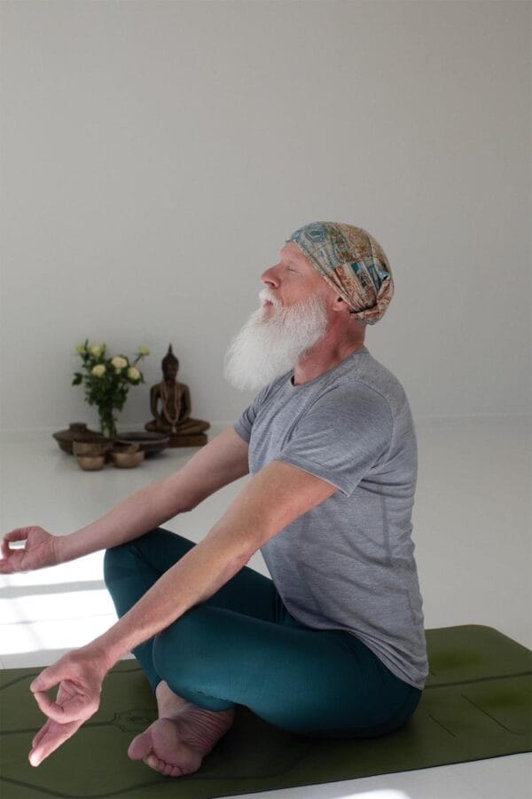 g21020_210_image_1 En gammel mand med hvidt skæg mediterer siddende på en yogamåtte, iført en farverig hovedbeklædning. I baggrunden er der en Buddha-statue og blomster i en vase.