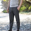 En person iført grå t-shirt og mørkegrå joggingbukser med hænderne i lommerne står på en grusvej omgivet af træer. Tekst på bukserne: "BEHOGEN".