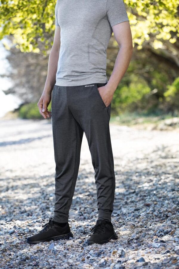 g21020_210_image_2 En person iført grå t-shirt og mørkegrå joggingbukser med hænderne i lommerne står på en grusvej omgivet af træer. Tekst på bukserne: "BEHOGEN".